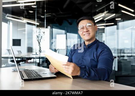 Ritratto di un uomo d'affari di successo all'interno di un moderno edificio di uffici, l'uomo asiatico ha ricevuto buone notizie in busta apre lettera felice sorridendo e guardando la fotocamera. Foto Stock