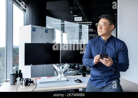 Ritratto di serio architetto asiatico uomo d'affari, uomo che guarda fuori finestra di lavoro in moderno luminoso ufficio interno utilizzando doppio computer, uomo d'affari tenendo il telefono in mano. Foto Stock
