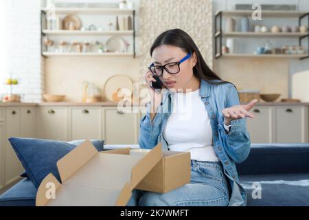 Donna insoddisfatta ha ricevuto un pacco rotto consegnato da un negozio online, litigi asiatici con il servizio di assistenza clienti, parla con l'operatore al telefono, seduto a casa in cucina. Foto Stock