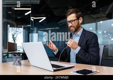 Videochiama il capo di un uomo d'affari di successo barba in tuta d'affari durante una riunione online, parlando con i colleghi utilizzando cuffie e computer portatile, per una conversazione remota, un uomo che lavora all'interno di un moderno edificio di uffici. Foto Stock