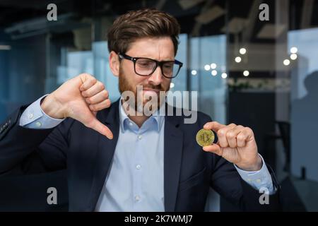 Foto ravvicinata di un uomo d'affari maschio che tiene in mano moneta bitcoin di valuta cripto oro, investitore maturo deluso con il tasso di cambio e l'investimento, capo in tuta di affari che mostra il dito giù Foto Stock