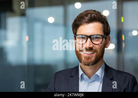 Primo piano ritratto fotografico di un uomo d'affari felice e di successo, boss maturo con barba e occhiali che lavorano all'interno di un moderno edificio per uffici, investitore senior sorridente e guardando la fotocamera. Foto Stock