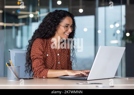 Giovane e felice donna ispanica che lavora in un ufficio moderno utilizzando un computer portatile, donna d'affari sorridente e felice in occhiali e capelli ricci. Foto Stock