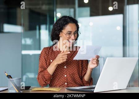 Businesswoman ha ricevuto felice lettera di notizie dalla banca, donna ispanica con capelli ricci e occhiali lavora all'interno di un moderno edificio ufficio, utilizza il computer portatile. Foto Stock