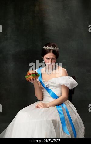 Tristezza. Giovane bella donna a immagine di regina o principessa in vestito bianco medievale con grande hamburger su sfondo scuro. Concetto di confronto di Foto Stock