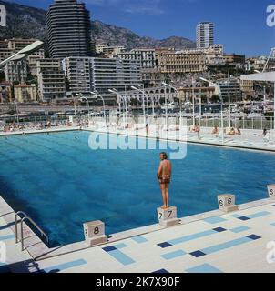 1964, storico, una vista dello Stato Nautique Rainier III, una piscina nel mezzo di Port Hercule a Monaco, con una piattaforma di immersione principale (non in foto) e più piccole, piattaforme in pietra lato piscina, una delle quali un nuotatore maschile è in piedi. La piscina olimpionica all'aperto di acqua di mare è stata ufficialmente aperta nell'agosto 1961. Foto Stock