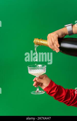 Sfondo astratto della festa di Natale, lo Champagne viene versato da una bottiglia in un bicchiere in una mano femminile. Minimalismo, sfondo verde, Natale A. Foto Stock