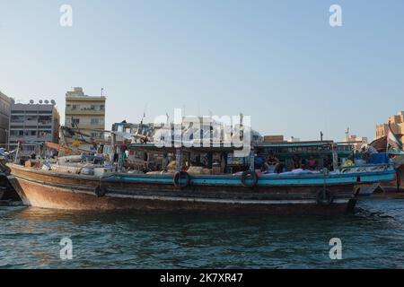 Vecchia e tradizionale nave da carico, chiamata un Dhow, ormeggiata al Dubai Creek, edifici di Deira dietro, a Dubai, Emirati Arabi Uniti. Gli uomini appendono fuori sul lato della barca. Foto Stock