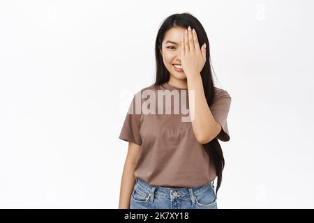 Ritratto di giovane donna asiatica coprire un lato del viso, guardando con un occhio, metà coperta di palma, sorridendo, controllando la visione al negozio ottico, bianco Foto Stock