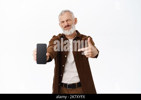 Vecchio deluso, anziano che mostra una cattiva app, che indica lo schermo dello smartphone, che si trova dispiaciuto contro lo sfondo bianco Foto Stock