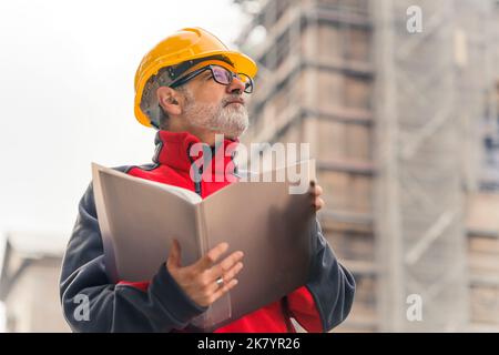Operaio professionista impegnato, caucasico, bearded, grigio-haired della costruzione in uniforme che indossa un casco arancione luminoso e gli occhiali, che tiene una cartella con i documenti, che guarda la macchina fotografica. Lavori di costruzione. Foto di alta qualità Foto Stock