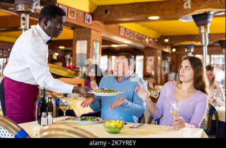 Dispiaciuta coppia arrabbiata seduta al tavolo al ristorante, parlando con confuso cameriere afroamericano Foto Stock