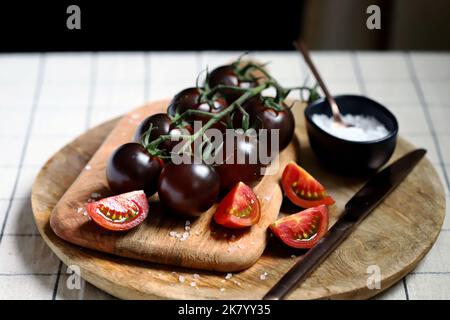 Pomodori neri su un ramo su un asse di legno con sale marino. Foto Stock