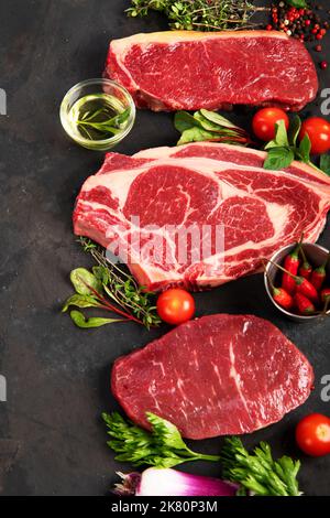 Varietà di bistecche di carne di manzo crudo per grigliare con condimento e utensili su sfondo scuro, vista dall'alto. Foto Stock