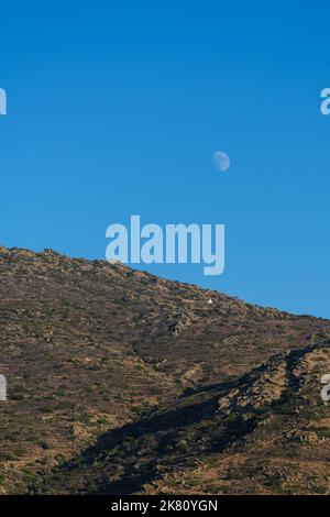 Vista di una montagna con una singola cappella bianca e la luna nel cielo blu in Grecia iOS Foto Stock