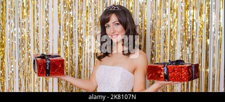 Bella bruna modello si trova su sfondo dorato e sorride bel sorriso, lei tiene un regalo in un abito bianco con un diadema sulla sua testa buon Natale e felice anno nuovo Foto Stock