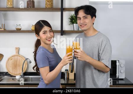 Giovane uomo in vestito casual e la sua ragazza bere succo di frutta misto e veggie frullato. Gli amanti asiatici trascorrono insieme il tempo della mattina in cucina. Foto Stock