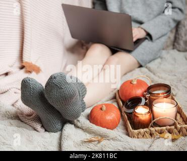 Una ragazza in un maglione e calzini a maglia si siede su una coperta e lavora su un computer portatile. Accanto a lei c'è un vassoio con tutte le cose e le zucche. Accogliente concetto autunnale. Foto Stock