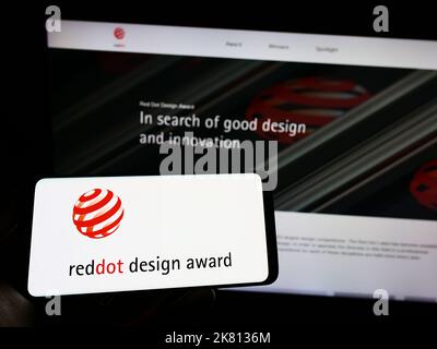 Persona che tiene il cellulare con il logo del premio internazionale tedesco Red Dot Design Award sullo schermo di fronte alla pagina web. Messa a fuoco sul display del telefono. Foto Stock
