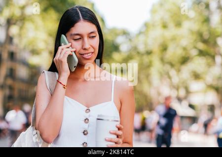 Donna ispanica positiva con una tazza di caffè ecocompatibile per sorridere e parlare con lo smartphone mentre si passa il tempo nel parco illuminato il giorno del fine settimana estivo a Barcellona, Spagna Foto Stock