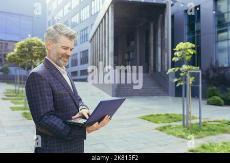 Boss felice e sorridente che usa il laptop mentre si trova fuori dal moderno edificio degli uffici, uomo d'affari maturo in tuta aziendale, investitore senior soddisfatto del risultato lavorativo. Foto Stock