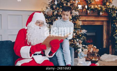 Babbo Natale sta leggendo il libro al ragazzo piccolo felice che si congratula con il bambino il giorno di Natale seduto sul divano in casa decorata. Concetto di infanzia e letteratura. Foto Stock