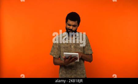 giovane afroamericano perplesso con barba con tavoletta digitale su sfondo arancione, immagine di scorta Foto Stock