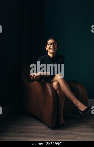 Donna asiatica sorridente sorridente alla macchina fotografica mentre si siede su una poltrona. Felice giovane donna che si rilassa nel suo ufficio. Foto Stock