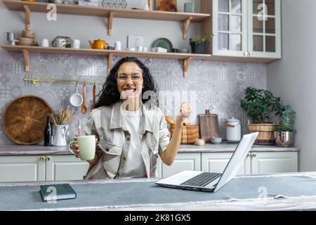 Ritratto di donna di successo a casa, donna ispanica guardando la macchina fotografica e felice celebrando trionfo vittoria, donna d'affari che utilizza il portatile per il lavoro remoto, freelance con capelli ricci e occhiali. Foto Stock