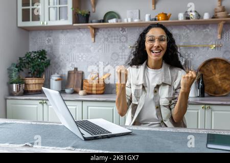 Ritratto di donna di successo a casa, donna ispanica guardando la macchina fotografica e felice celebrando trionfo vittoria, donna d'affari che utilizza il portatile per il lavoro remoto, freelance con capelli ricci e occhiali. Foto Stock