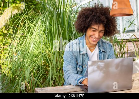 Giovane ragazzo sorridente di etnia 20 anni, vestito in abiti casual, lavora su un computer portatile all'aperto, in una zona verde in natura. Spazio verde aperto per Foto Stock