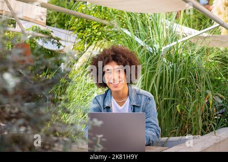 Giovane ragazzo sorridente di etnia 20 anni, vestito in abiti casual, lavora su un computer portatile all'aperto, in una zona verde in natura. Spazio verde aperto per Foto Stock