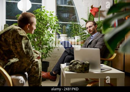 Giovane donna afflitta in uniforme militare che parla con psicologo durante la sessione di terapia, al chiuso. Aiuto, supporto, ptsd, salute e concetto di armonia Foto Stock