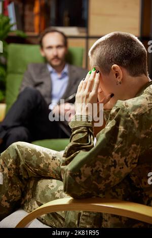 Giovane donna afflitta in uniforme militare che parla con psicologo durante la sessione di terapia, al chiuso. Aiuto, supporto, ptsd, salute e concetto di armonia Foto Stock