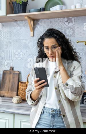 Colpo verticale, donna triste a casa leggendo cattive notizie sul telefono, donna ispanica a casa con capelli ricci e occhiali depressi tenendo smartphone. Foto Stock