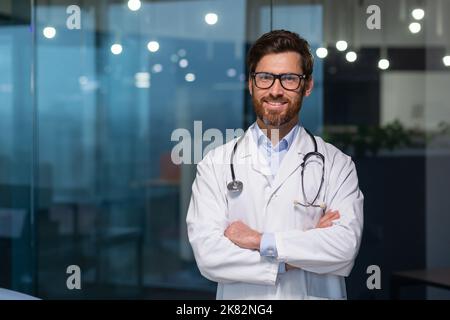 Ritratto di medico maturo con barba, uomo in camice medico bianco sorridente e guardando la macchina fotografica a braccia incrociate lavorando all'interno di clinica moderna. Foto Stock