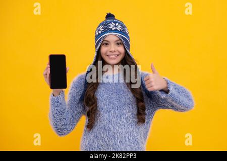 Bambino adolescente in inverno cappello a maglia che mostra schermo vuoto telefono cellulare, mock su spazio copia. Faccia felice, emozioni positive e sorridenti della ragazza adolescente Foto Stock