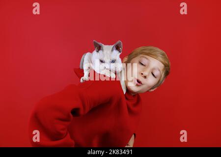 Un ragazzo allegro in tuta rossa con un piccolo gattino bianco con occhi blu seduti sulle spalle sale sulle spalle del ragazzo Foto Stock