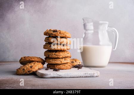 Gustosi biscotti fatti in casa con scaglie di cioccolato e caraffa di latte in vetro su fondo grigio in cemento Foto Stock