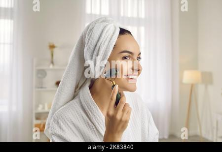 Giovane donna felice e sorridente che massaggia il suo bel viso con un rullo facciale in giada Foto Stock