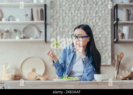 Ritratto di una giovane bella donna asiatica seduta in cucina al tavolo, tenendo una fresca insalata verde nella forchetta, sospirando, guardandola infamia. Stanco di stare, odia il vegetarismo. Foto Stock