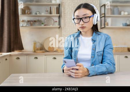 Giovane bella donna asiatica concentrata ascoltando audiolibro, podcast. Seduti a casa in cucina indossando cuffie bianche e tenendo il telefono. Foto Stock
