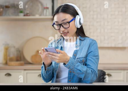 Giovane bella donna asiatica che ascolta musica al telefono. Seduti a casa in cucina indossando cuffie bianche, tenendo uno smartphone, scorrendo la playlist, scegliendo una canzone, sorridendo Foto Stock