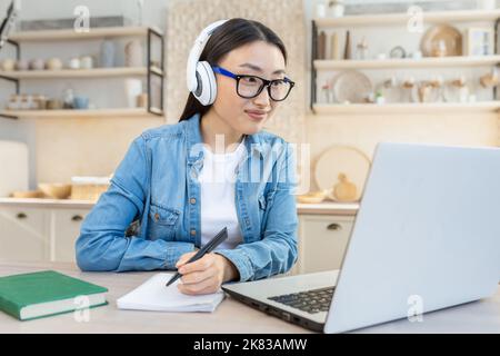 Formazione online. Giovane bella ragazza asiatica seduta al tavolo a casa in cuffie bianche e con un computer portatile. Studia in remoto. Scrive una lezione, una classe in un notebook. Foto Stock