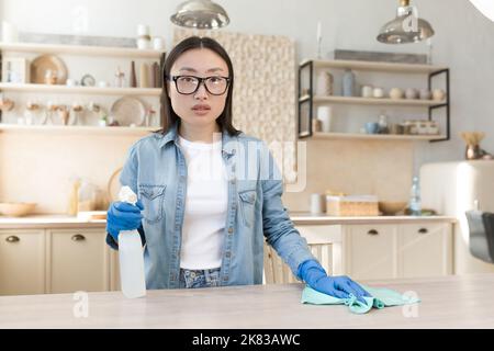 Ritratto di una giovane casalinga asiatica che pulisce a casa. Strofinare il tavolo in cucina con uno straccio in guanti di gomma, utilizza prodotti chimici per la casa. Sta in piedi, tenendo uno spray, guardando la telecamera. Foto Stock