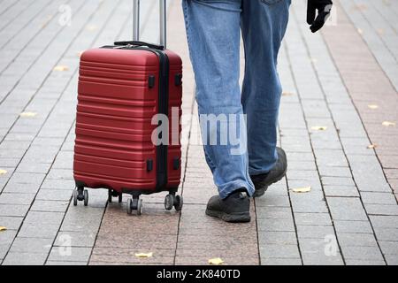 Uomo in jeans a piedi con valigia rossa su ruote. Gambe maschili e bagagli per strada, concetto di viaggio Foto Stock