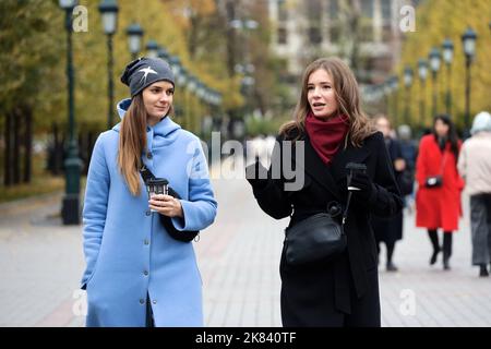 Due graziose ragazze in autunno vestiti che parlano mentre camminano sulla strada della città con le bevande del caffè, discussione emotiva Foto Stock