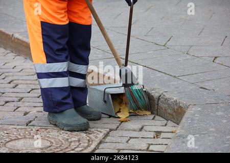 Operaio in uniforme con una scopa pulire il marciapiede, raccoglie le foglie cadute in una paletta. Pulizia della strada nella città d'autunno Foto Stock