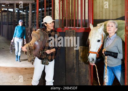 Donna anziana rancher che porta cavallo fuori stalla, mentre donna asiatica in sella Foto Stock