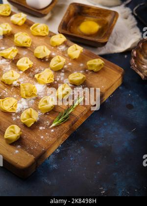 Su fondo di marmo blu scuro, un tagliere di legno cosparso di farina e gnocchi crudi con ripieno di carne e un rametto di rosmarino profumato. Re Foto Stock